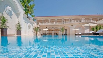 Una piscina con suelo de baldosas, rodeada de sillas de descanso y sombrillas, con un edificio y un balcón al fondo.