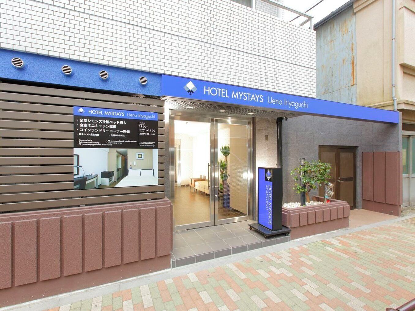 A hotel entrance with a blue awning, a reception desk, and a signboard displaying room rates and amenities.