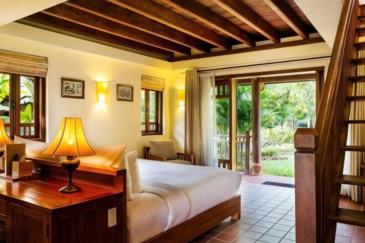 Quarto de hotel com cama, escrivaninha com luminária, cadeira e vista para o exterior.