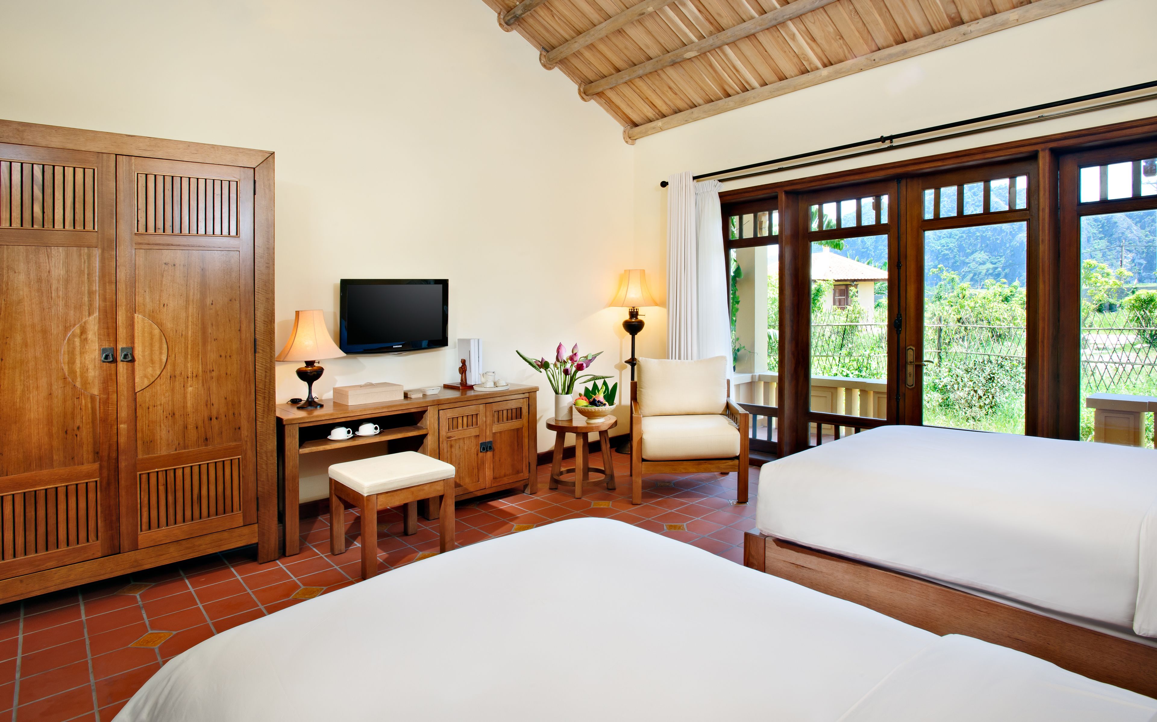 A hotel room with two beds, a TV, a desk with a lamp, a chair, and a view of the outdoors.