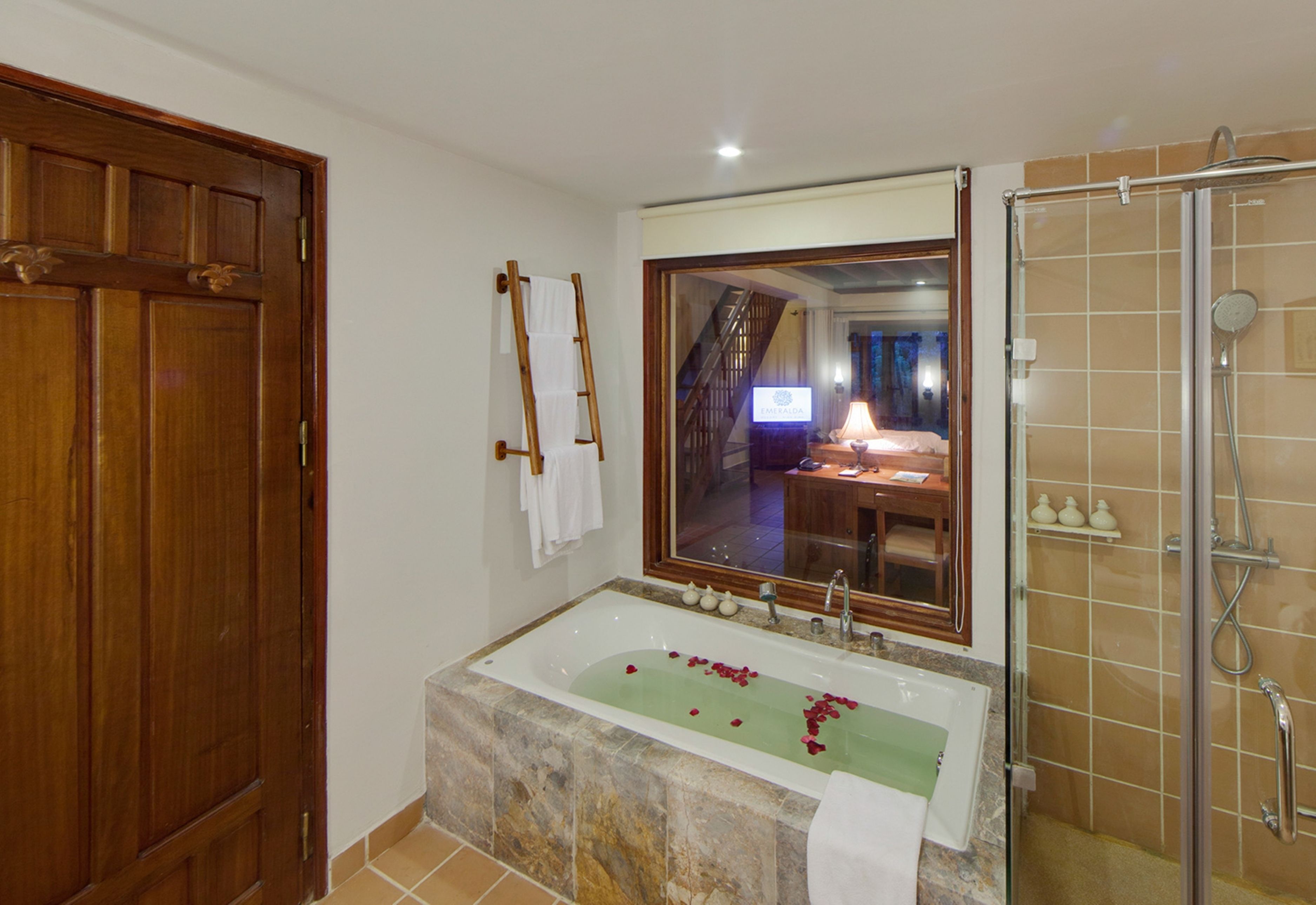 A bathroom with a large bathtub, a shower, a wooden door, and a mirror reflecting a room with a desk and a computer.