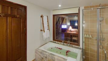 A bathroom with a large bathtub, a shower, a wooden door, and a mirror reflecting a room with a desk and a computer.