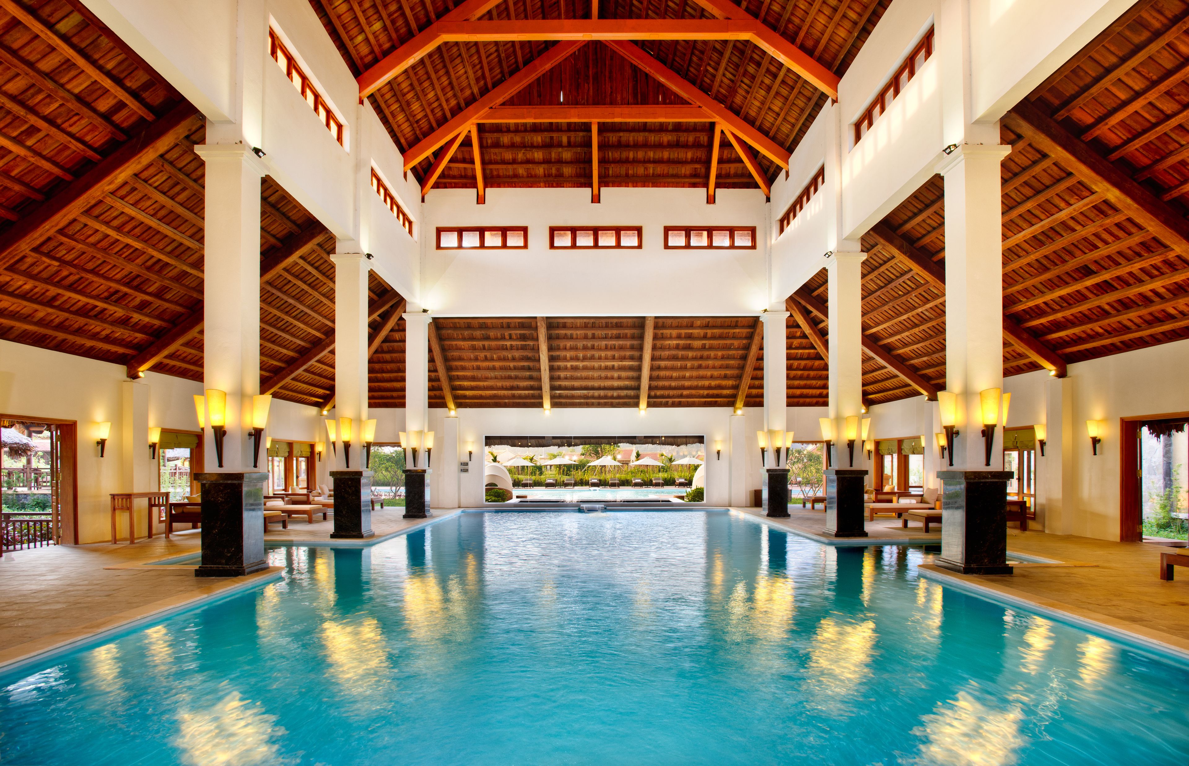 Una amplia piscina interior con techo de madera alto y columnas, rodeada de zonas de descanso y vistas al exterior.