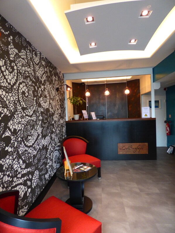 Un espace d’accueil avec un mur décoré de motifs, un comptoir de réception, un fauteuil rouge, une petite table avec des magazines et une plante en pot.