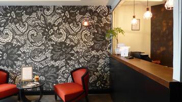 Zona de recepción con pared decorada, dos sillas rojas, una mesa redonda con un menú y un mostrador de recepción con una planta y un certificado enmarcado.