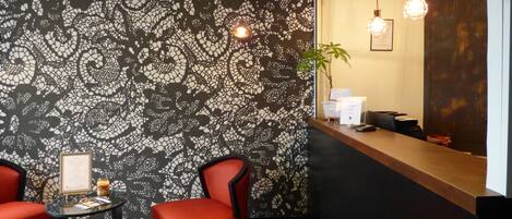 Zona de recepción con pared decorada, dos sillas rojas, una mesa redonda con un menú y un mostrador de recepción con una planta y un certificado enmarcado.