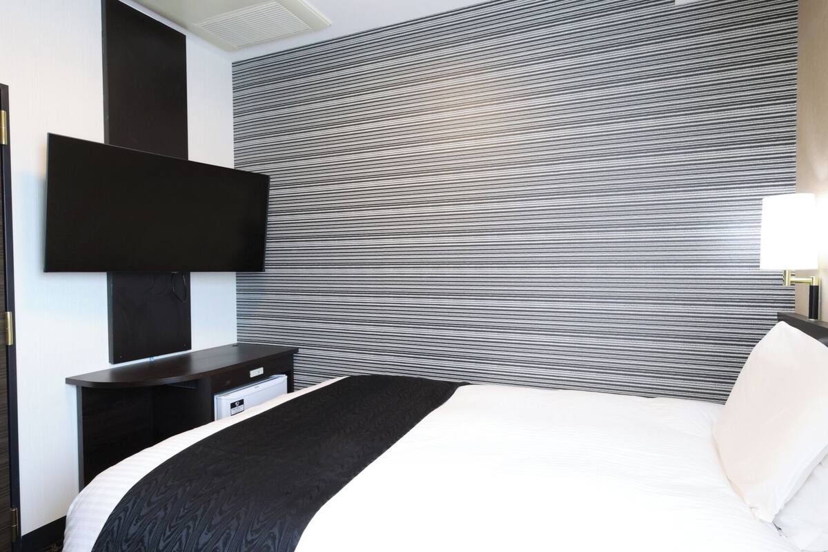 A hotel room with a bed, a flat-screen TV mounted on the wall, a bedside table with a lamp, and a striped wall design.