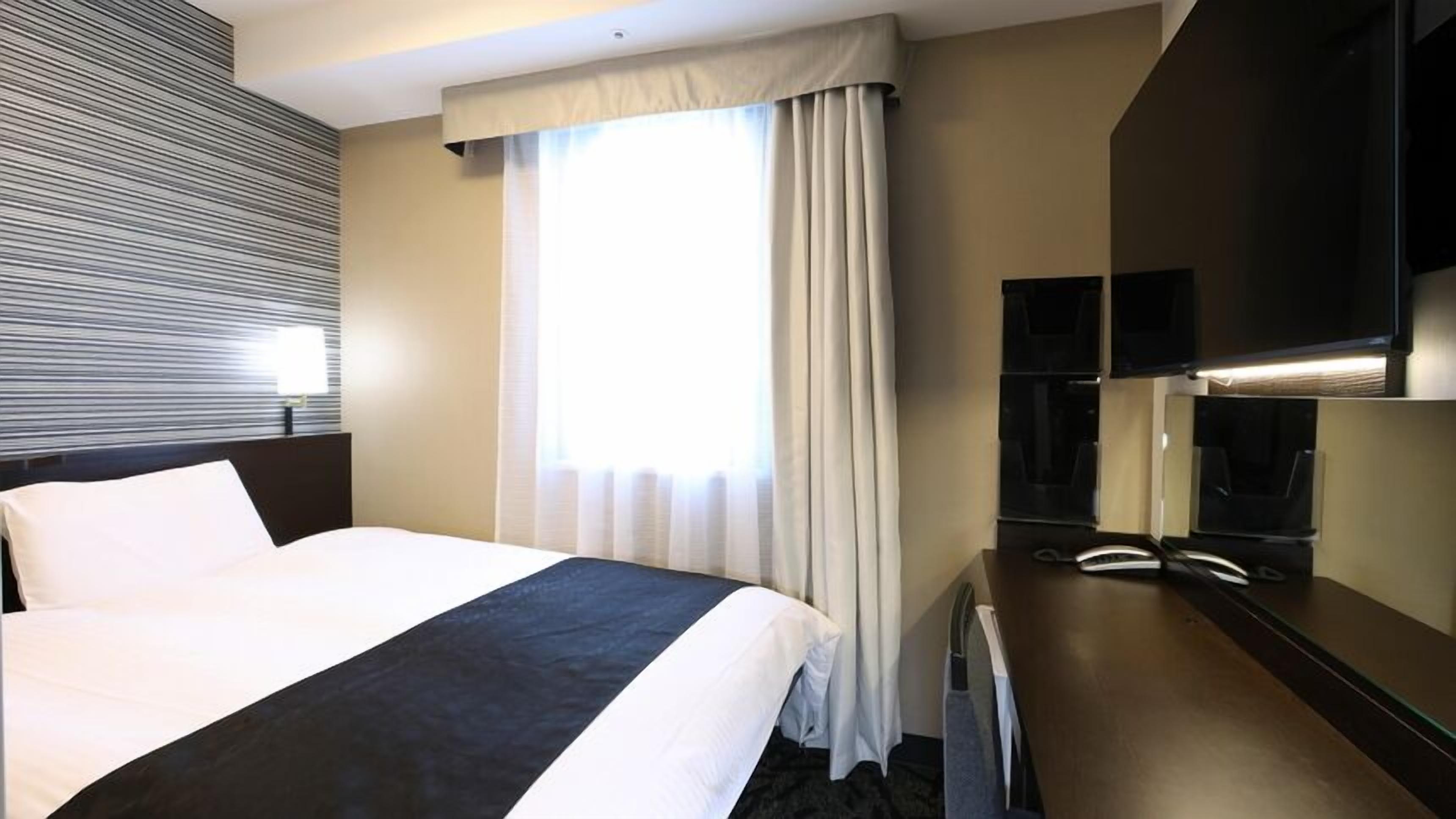 A hotel room with a bed, a desk with a telephone, a television, and a window with curtains.