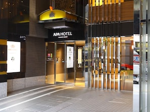 L'esterno dell'hotel APA HOTEL, dal design moderno, con un ingresso ben visibile caratterizzato da vetrate e un tendone giallo.