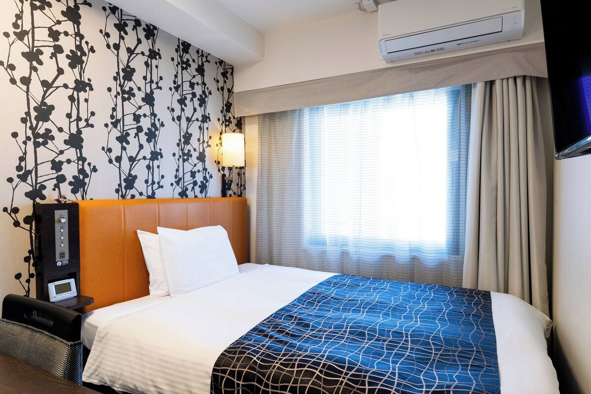 Ein Hotelzimmer mit einem Bett, einem Nachttisch mit Lampe, einer Klimaanlage und einem Fenster mit Vorhängen.
