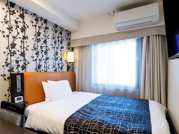 Habitación de hotel con cama, mesita de noche con lámpara, unidad de aire acondicionado y ventana con cortinas.