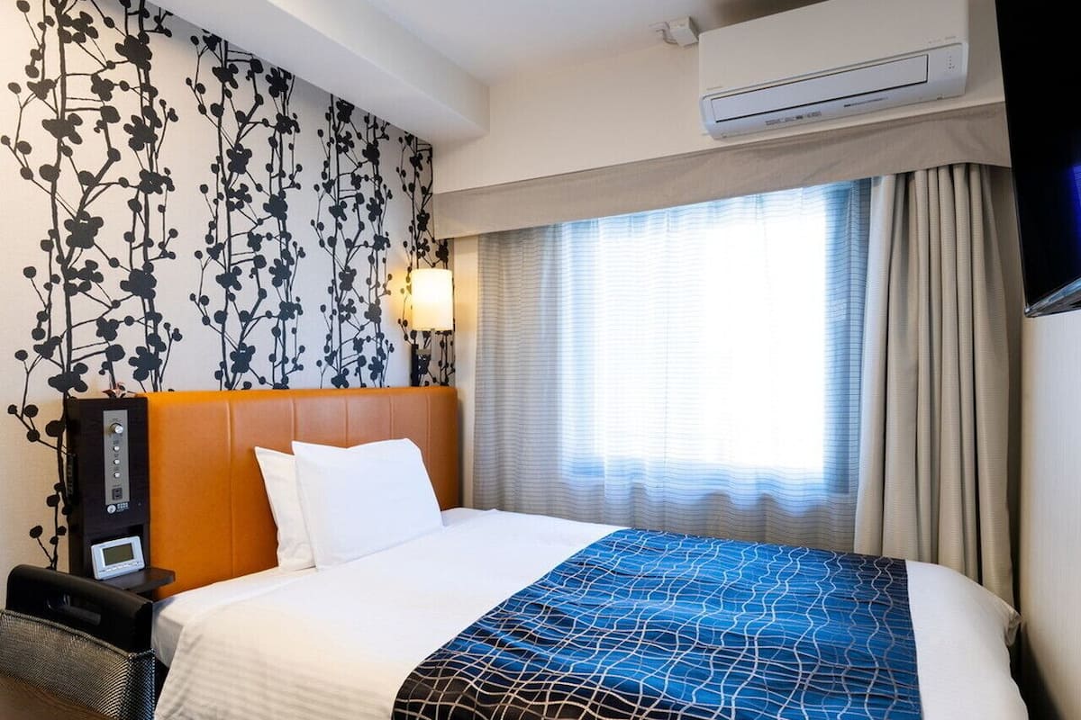 A hotel room with a bed, a nightstand, a wall-mounted air conditioner, and a window with curtains.
