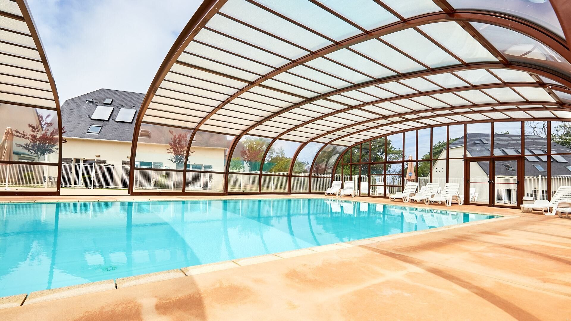 Una piscina con techo de vidrio, rodeada de sillas de descanso y una valla.