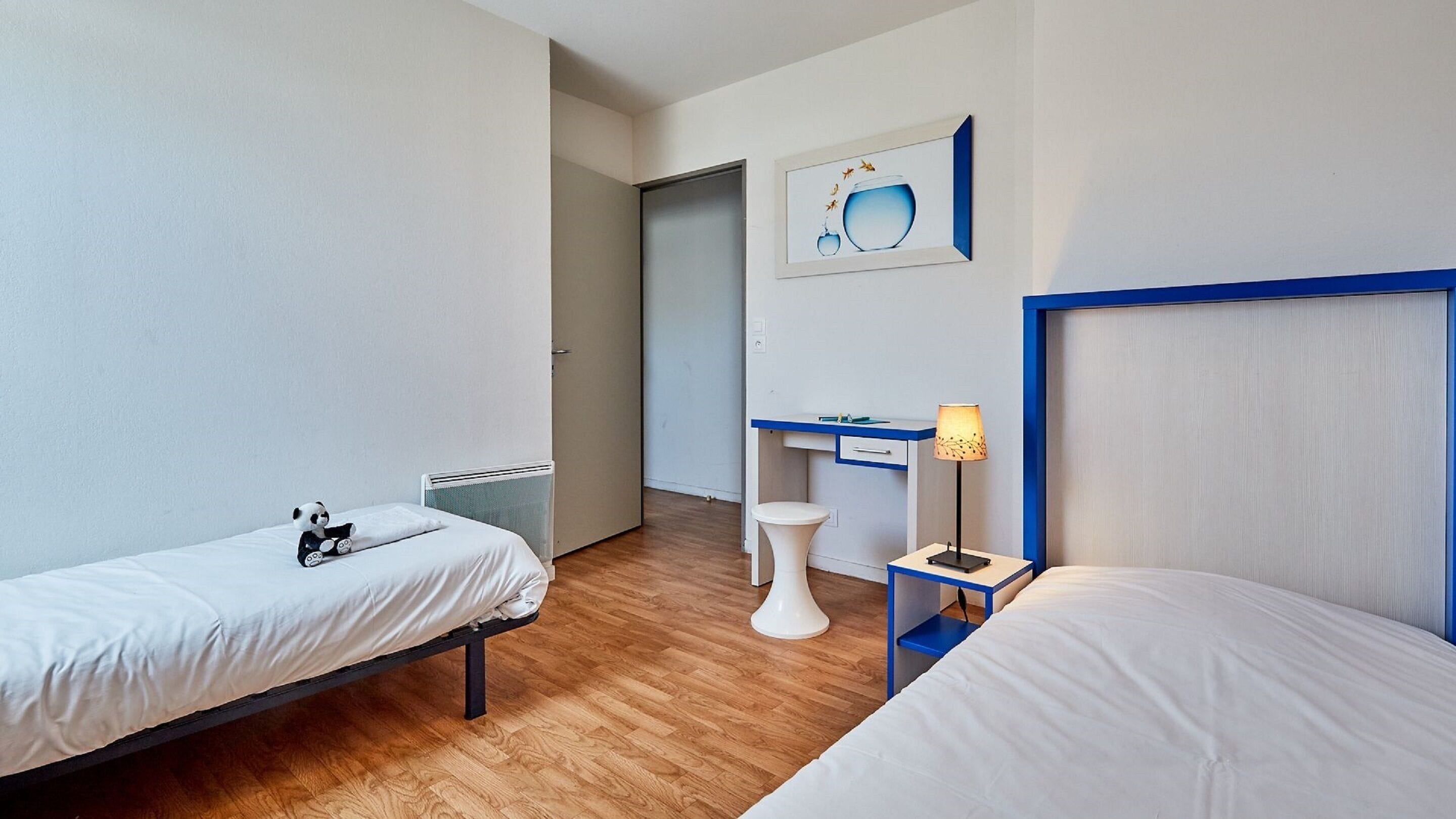 Une chambre d’hôtel avec deux lits, un bureau, une chaise, un radiateur et un tableau encadré au mur.