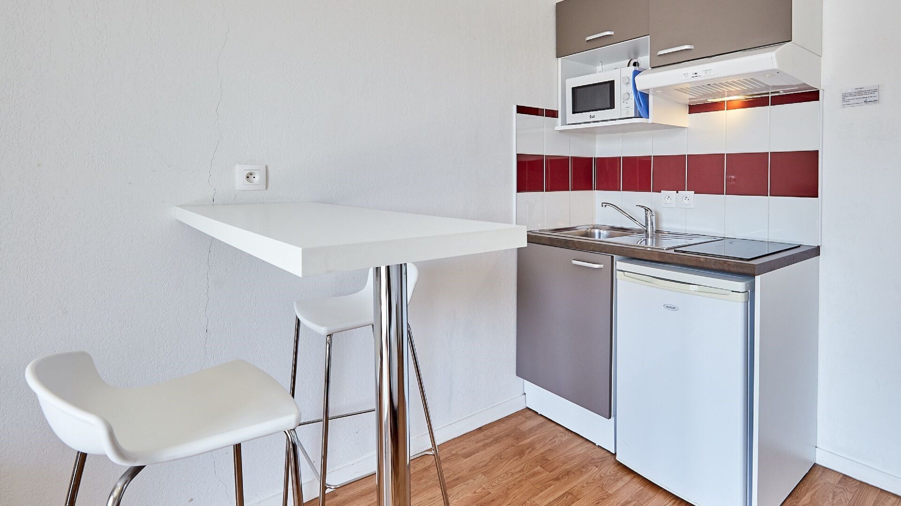 A compact kitchenette with a white countertop, a microwave, a sink, and a dishwasher. There are two white bar stools.