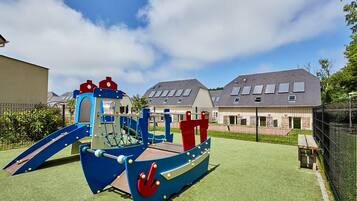 Children can play on the playground's slide and climbing elements on a well-maintained lawn surrounded by a fence.