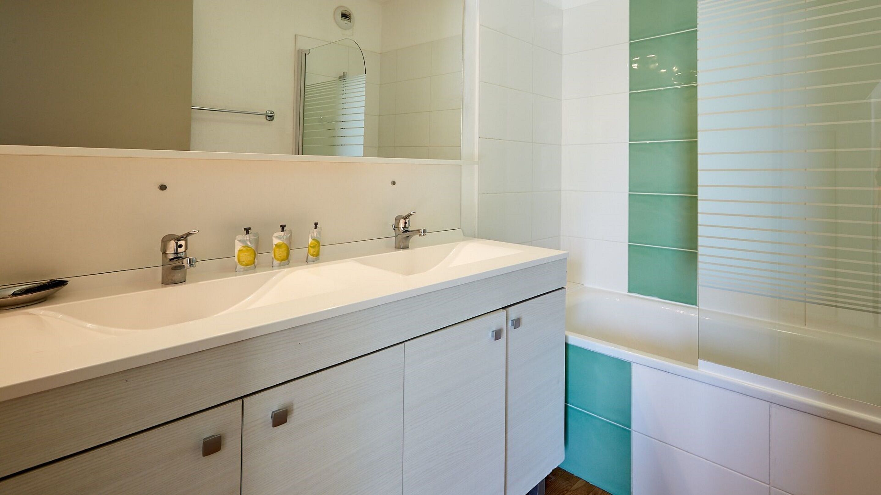 Une salle de bain moderne avec un meuble-lavabo blanc, un grand miroir et une baignoire recouverte de carreaux verts.