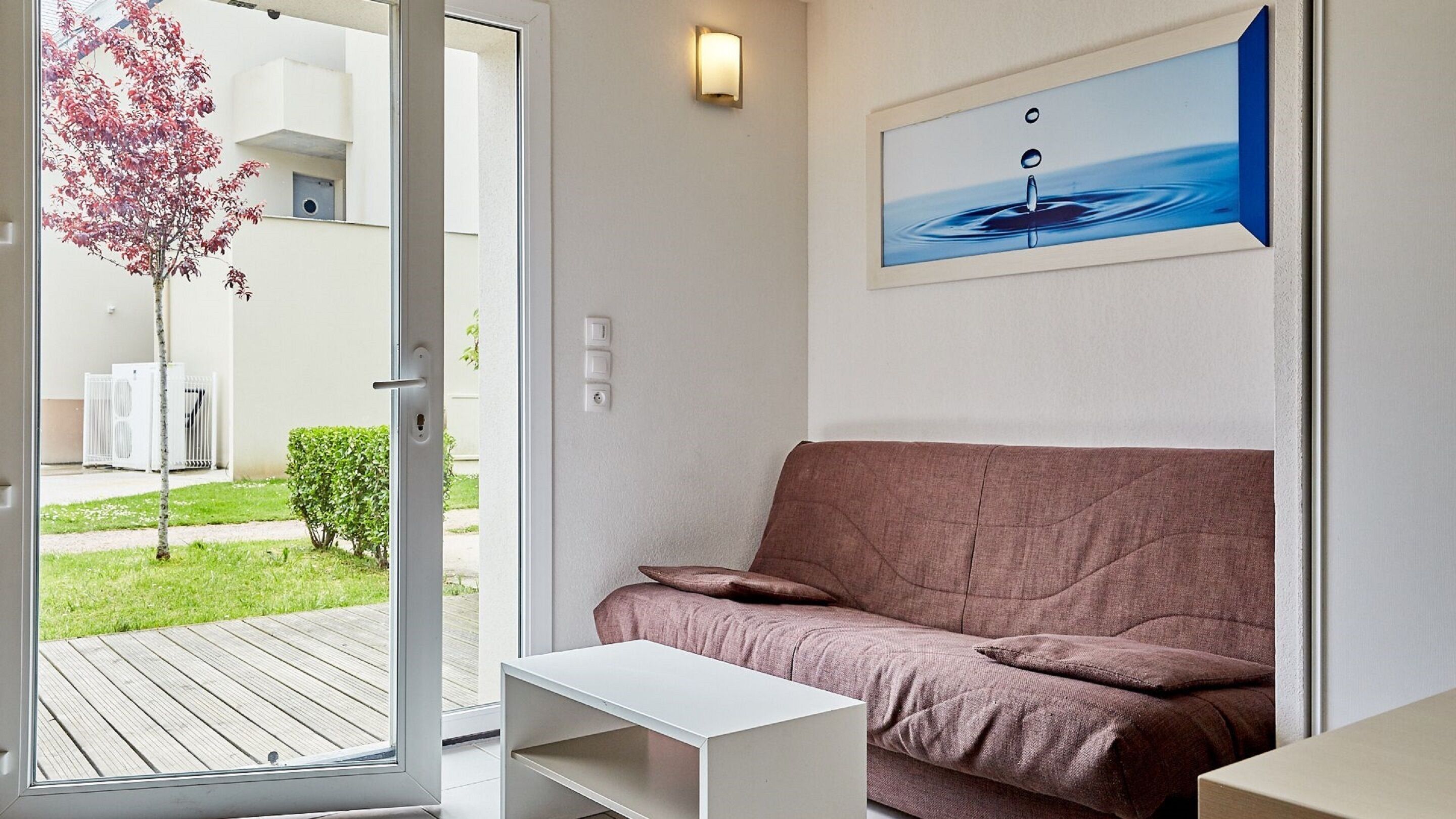 A modern living room with a brown sofa, a white coffee table, and a glass door leading to an outdoor area with a tree and a white building.