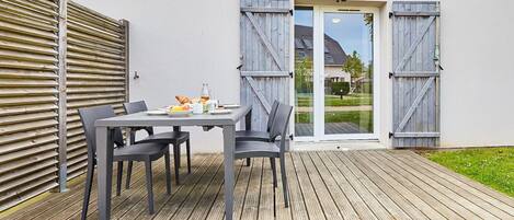 A patio with a table set for breakfast, including croissants and fruit, and a view of a house through a glass door.