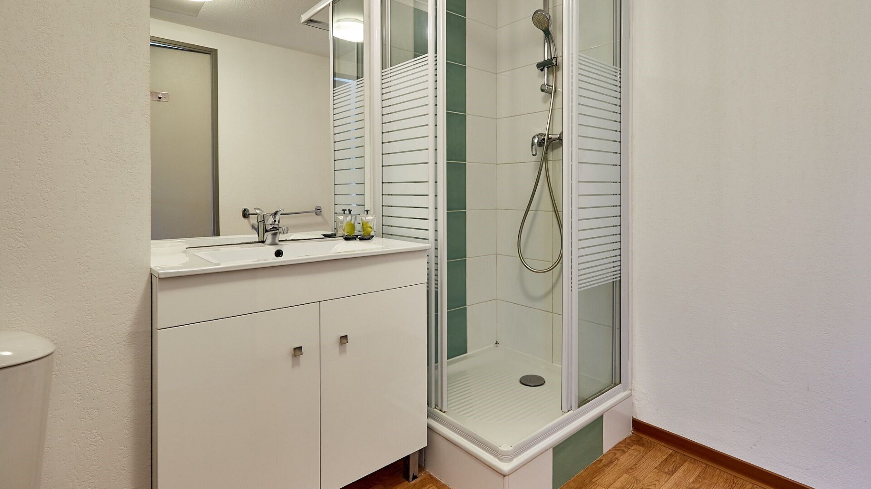 A modern bathroom with a glass-enclosed shower, a white sink with a mirror, and a wooden floor.