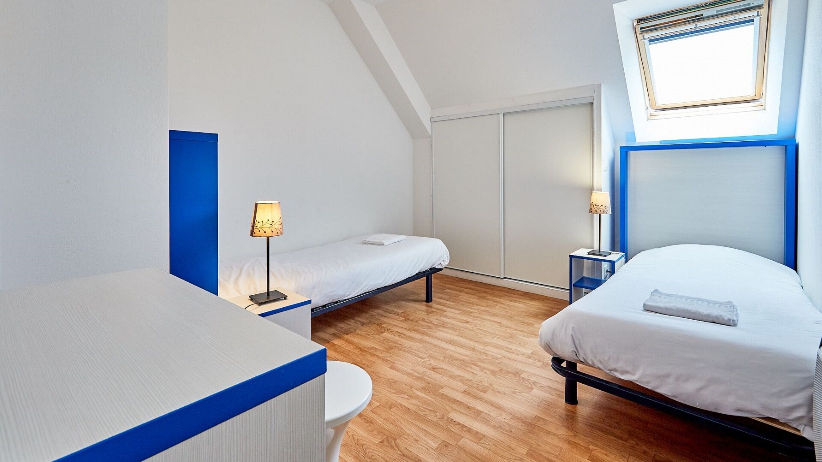 Een moderne hotelkamer met een open plafond, twee enkele bedden, een bureau en een kleintje eettafeltje.