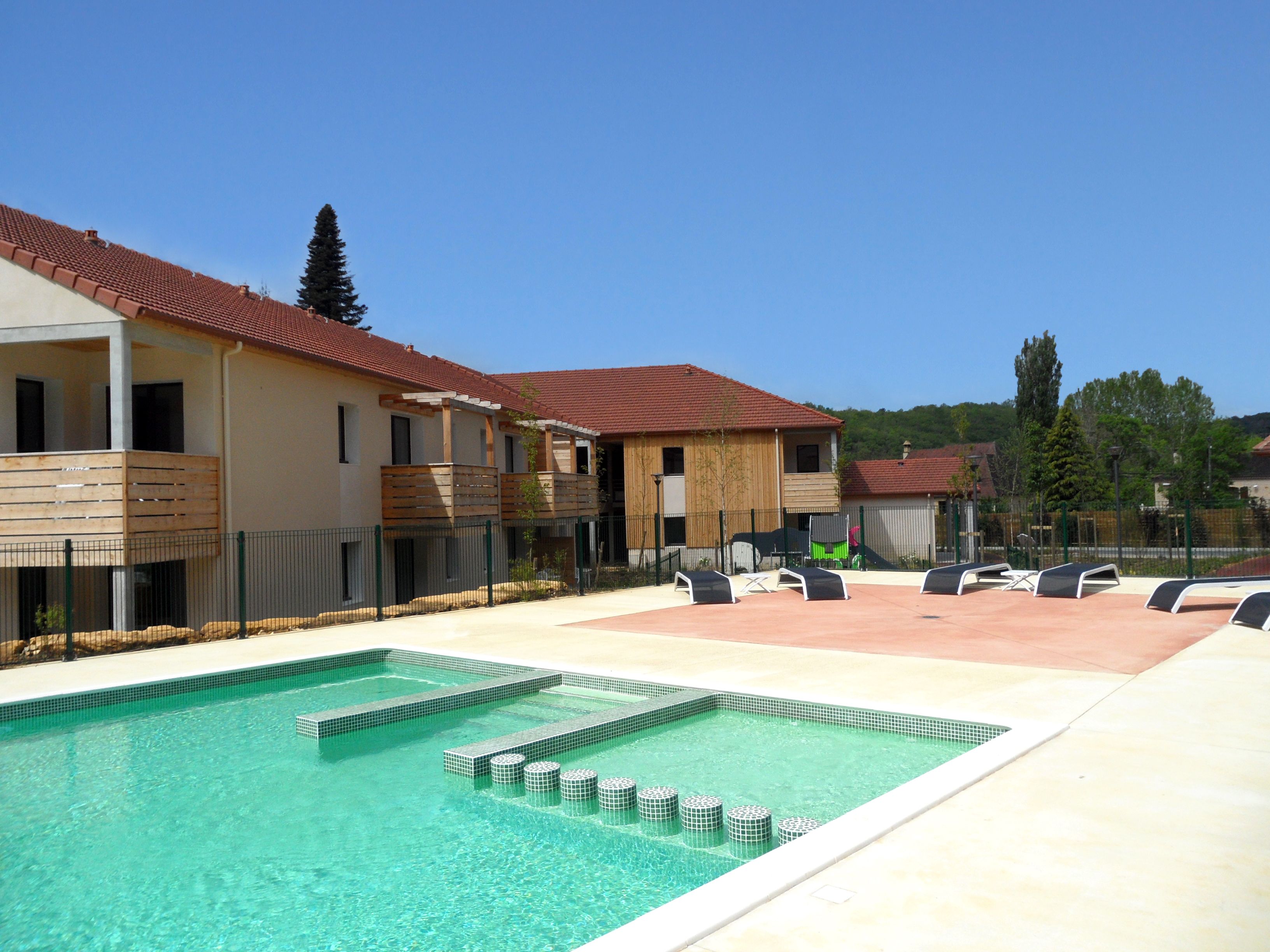 A swimming pool with a tiled deck, a covered seating area, and a building with a red-tiled roof.