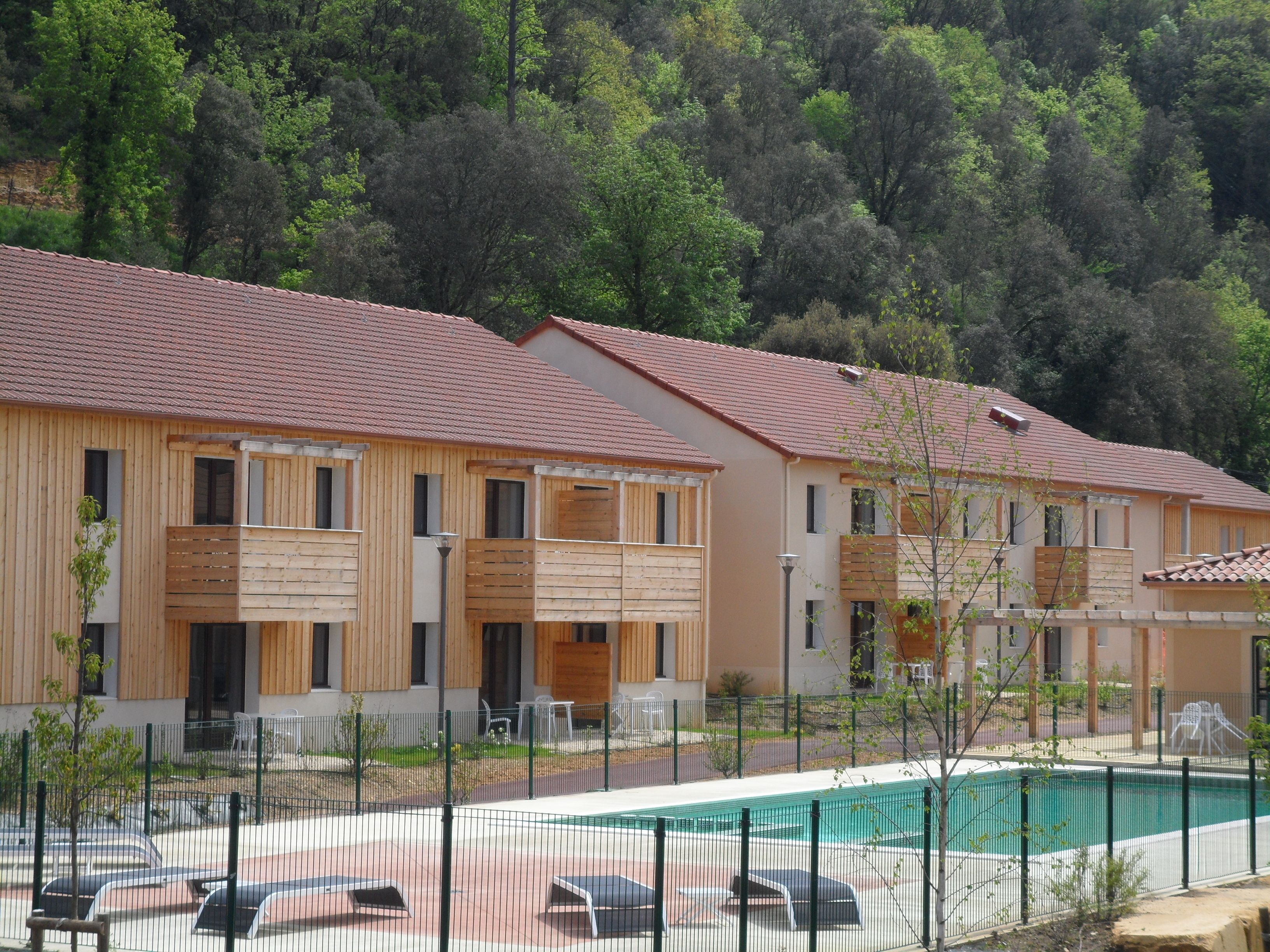 A row of wooden buildings with balconies, a swimming pool, and a forested hillside in the background.