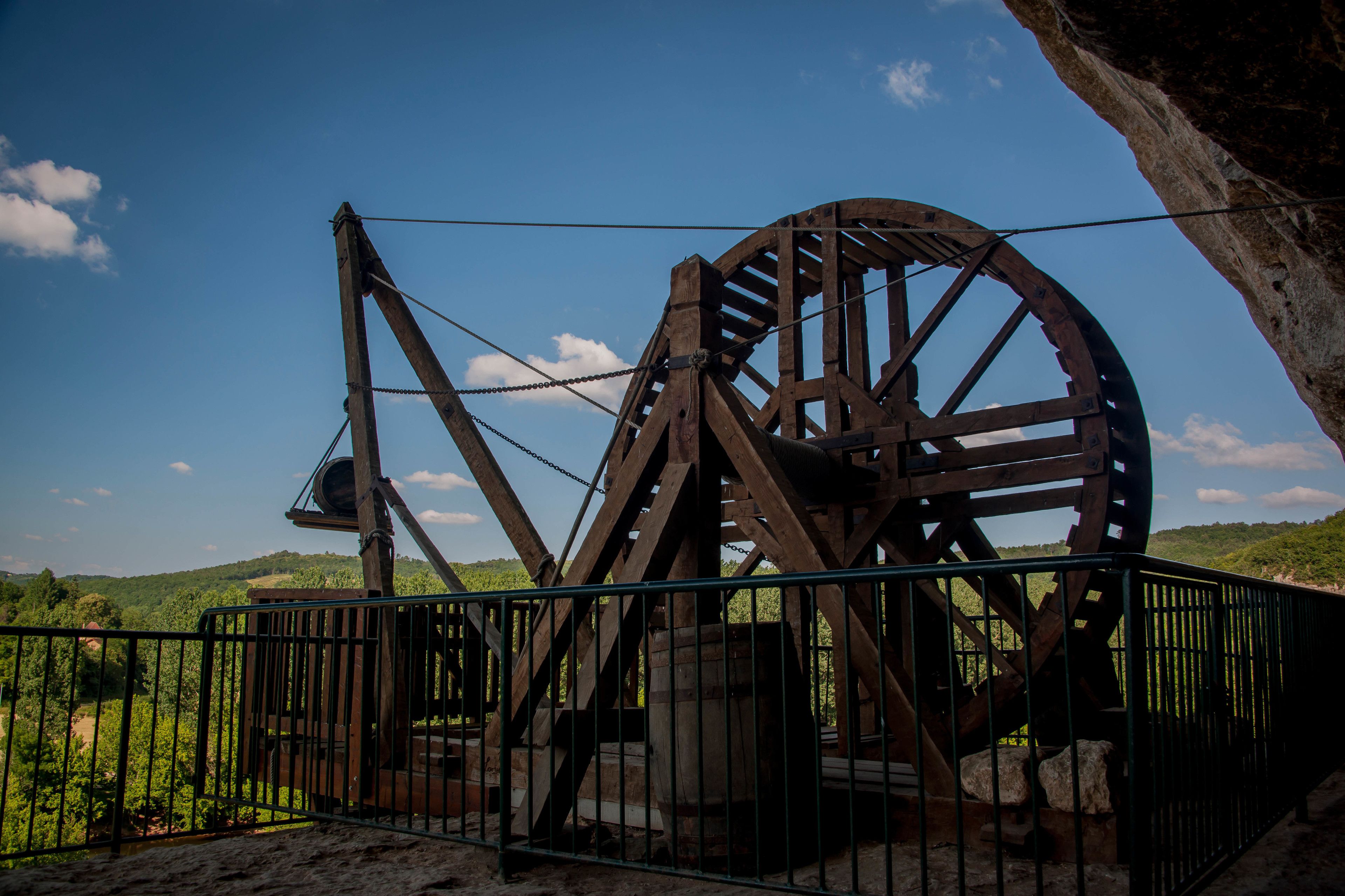 A large wooden water wheel structure with metal beams and chains, surrounded by a metal fence, set against a backdrop of green hills and a clear blue sky.