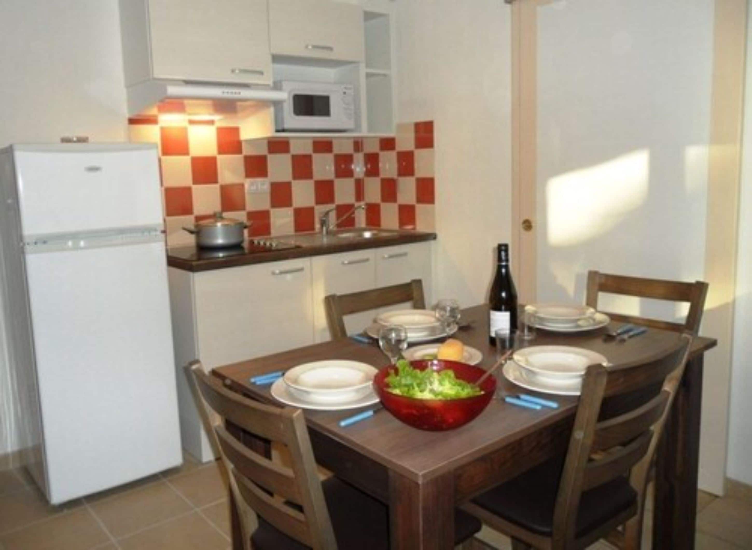 A compact kitchen with a microwave, a refrigerator, and a checkered backsplash. There is a dining table set with plates, glasses, and a bottle of wine.
