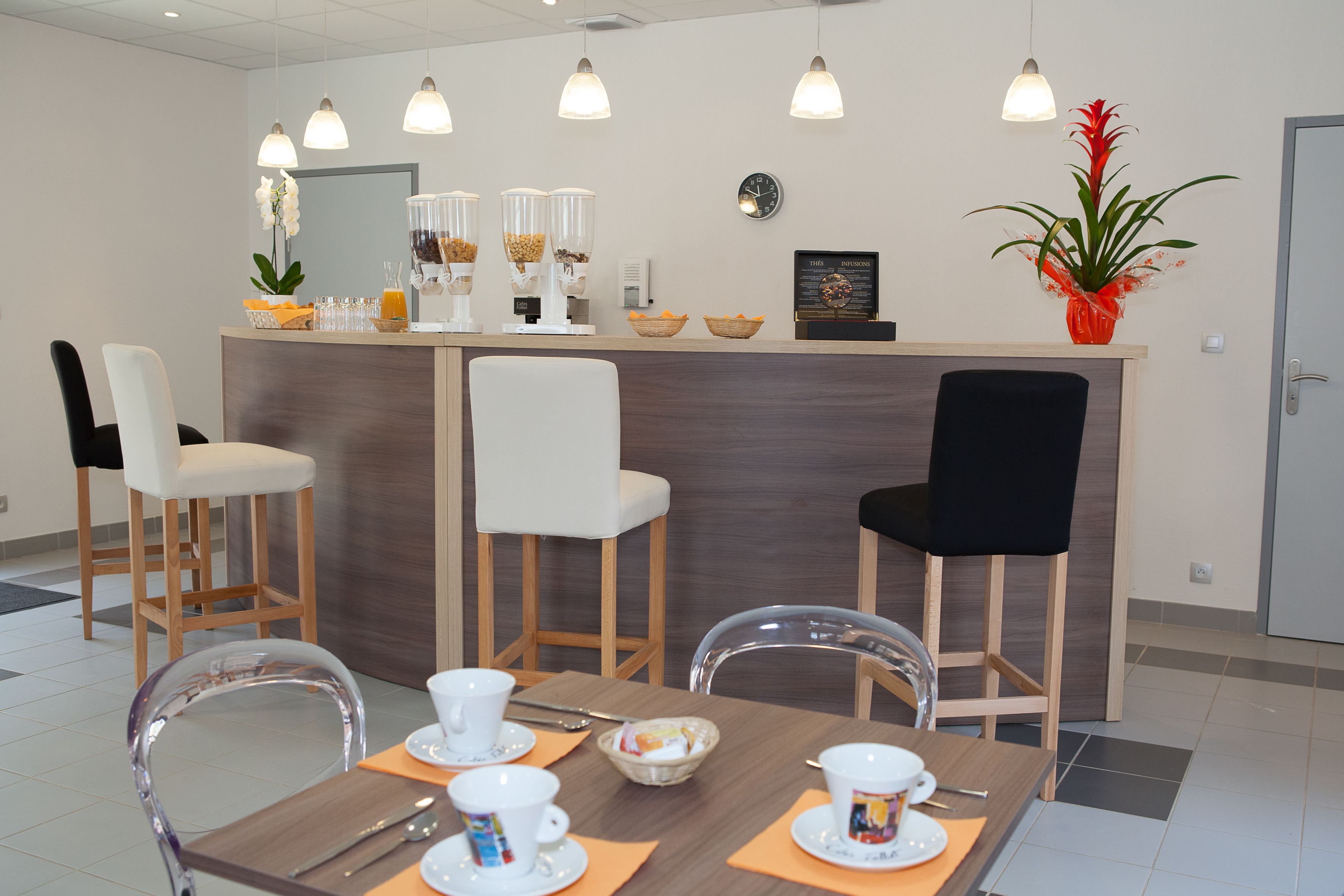 Zona de recepción de hotel con mostrador de madera, sillas blancas y negras, una mesa de comedor con alimentos para el desayuno y una planta en maceta.