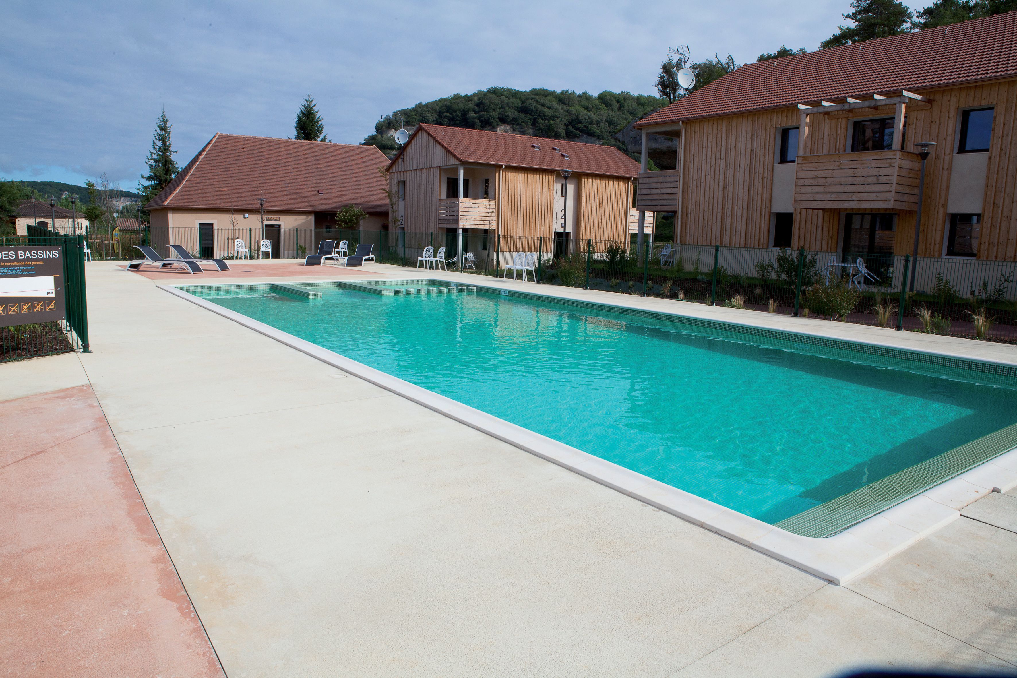 A rectangular swimming pool with a tiled deck, surrounded by wooden buildings with balconies.