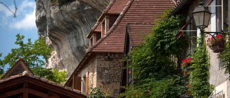 A traditional stone building with a steep roof, surrounded by lush greenery and a prominent rock formation in the background.