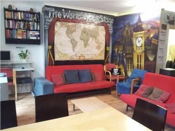 A cozy living room with a red sofa, blue armchairs, a wooden coffee table, and a bookshelf filled with books. A large world map mural and a clock tower image adorn the wall. - Central Hotel - Hostel (London)