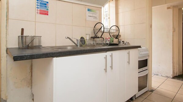 A kitchen with white cabinets, a stainless steel sink, and a dish rack. There are signs on the wall, including a fire action sign and a clear as you go sign. - Central Hotel - Hostel (London)