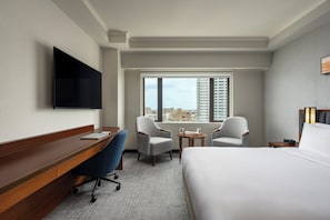 Habitación de hotel con una cama, un escritorio, dos sillones, una mesita, un televisor montado en la pared y una ventana con vistas a la ciudad.