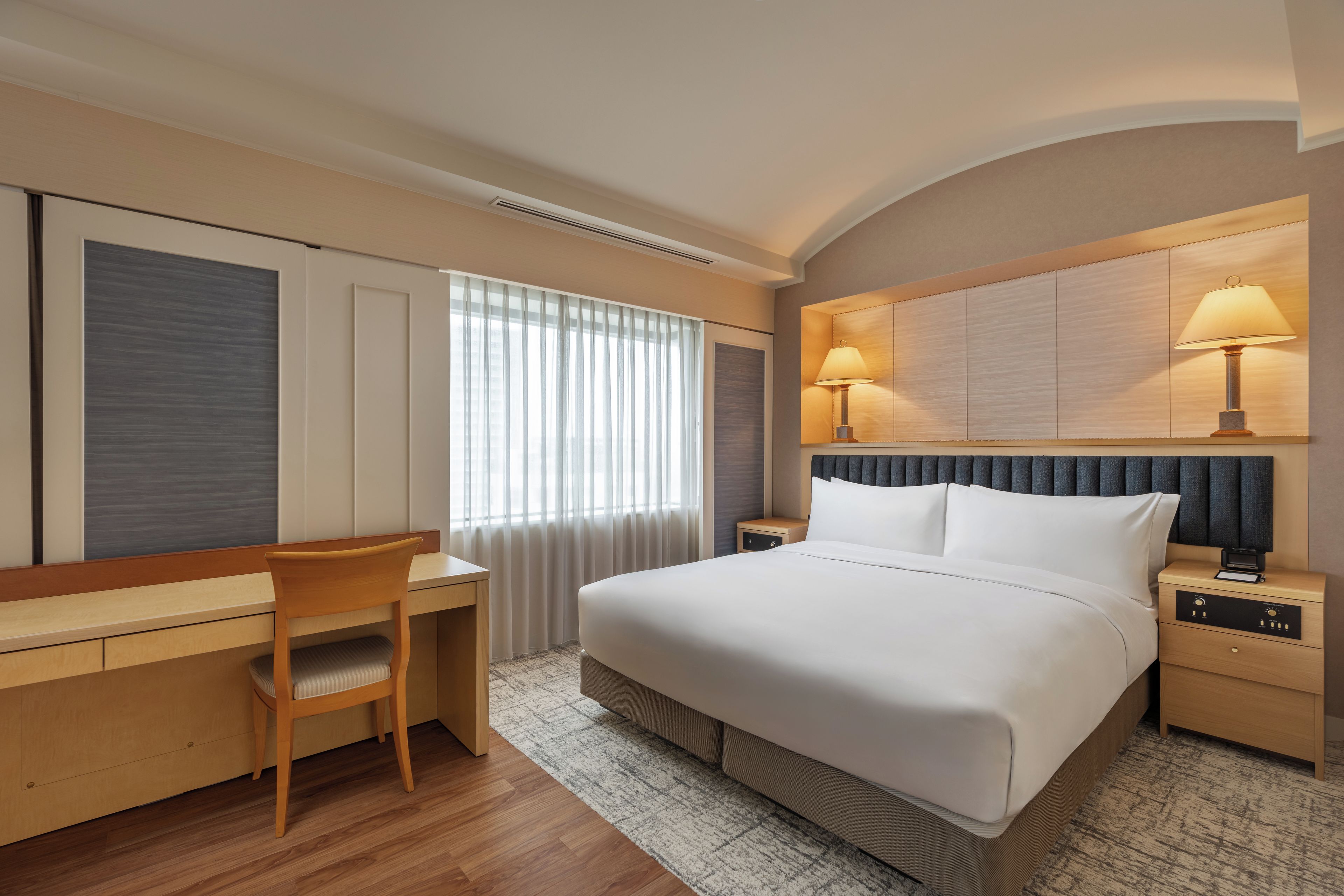 Ein Hotelzimmer mit Bett, Schreibtisch, Stuhl, Nachttischen, Lampen und einem fenster mit Vorhängen.