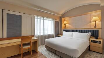 A hotel room with a bed, desk, chair, bedside tables, lamps, and a window with curtains.