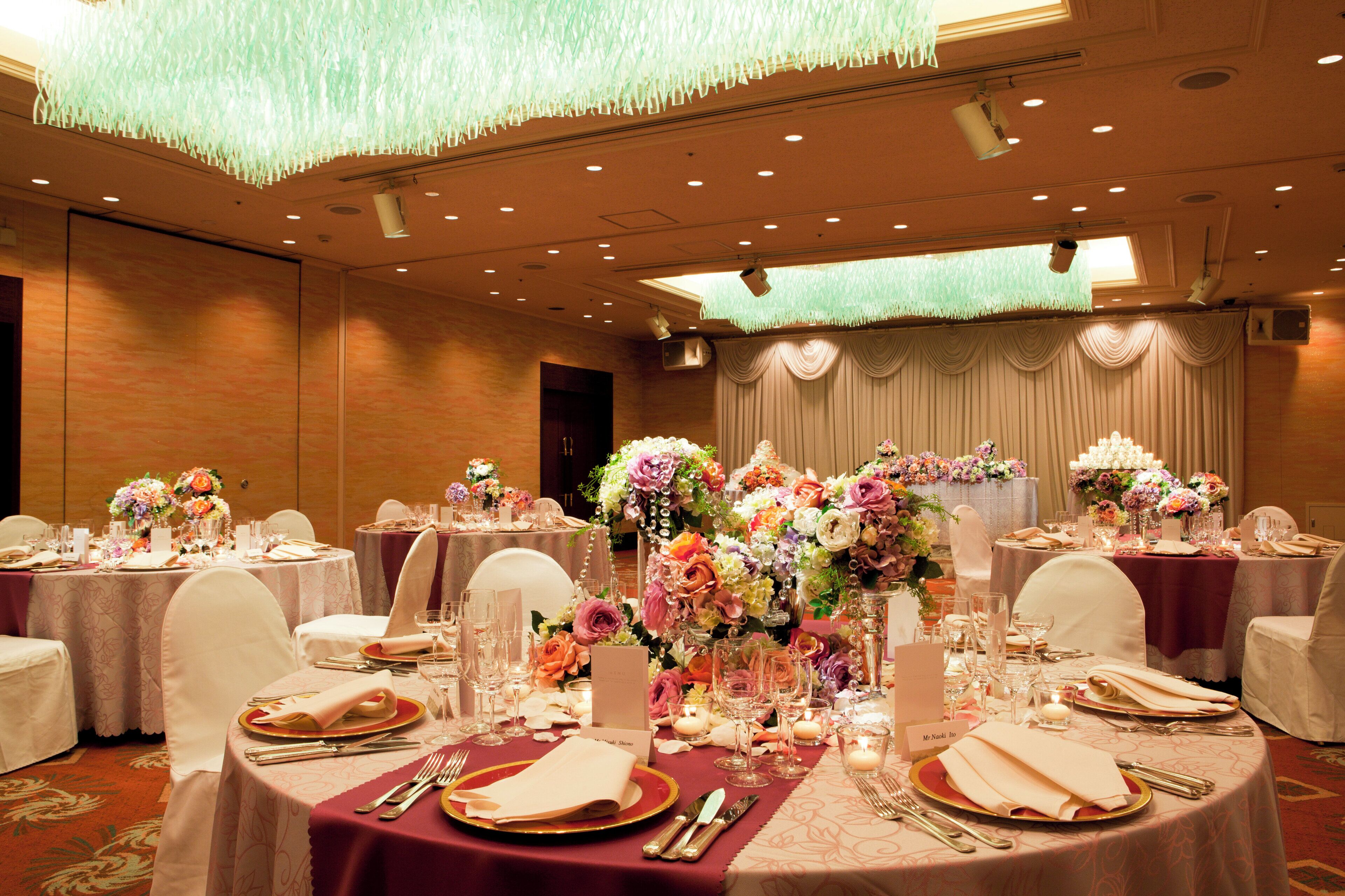 A banquet hall with round tables set for a formal event, featuring floral centerpieces and a chandelier.
