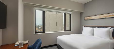 A hotel room with a bed, a desk, a chair, a window with a view of buildings, and a painting on the wall.
