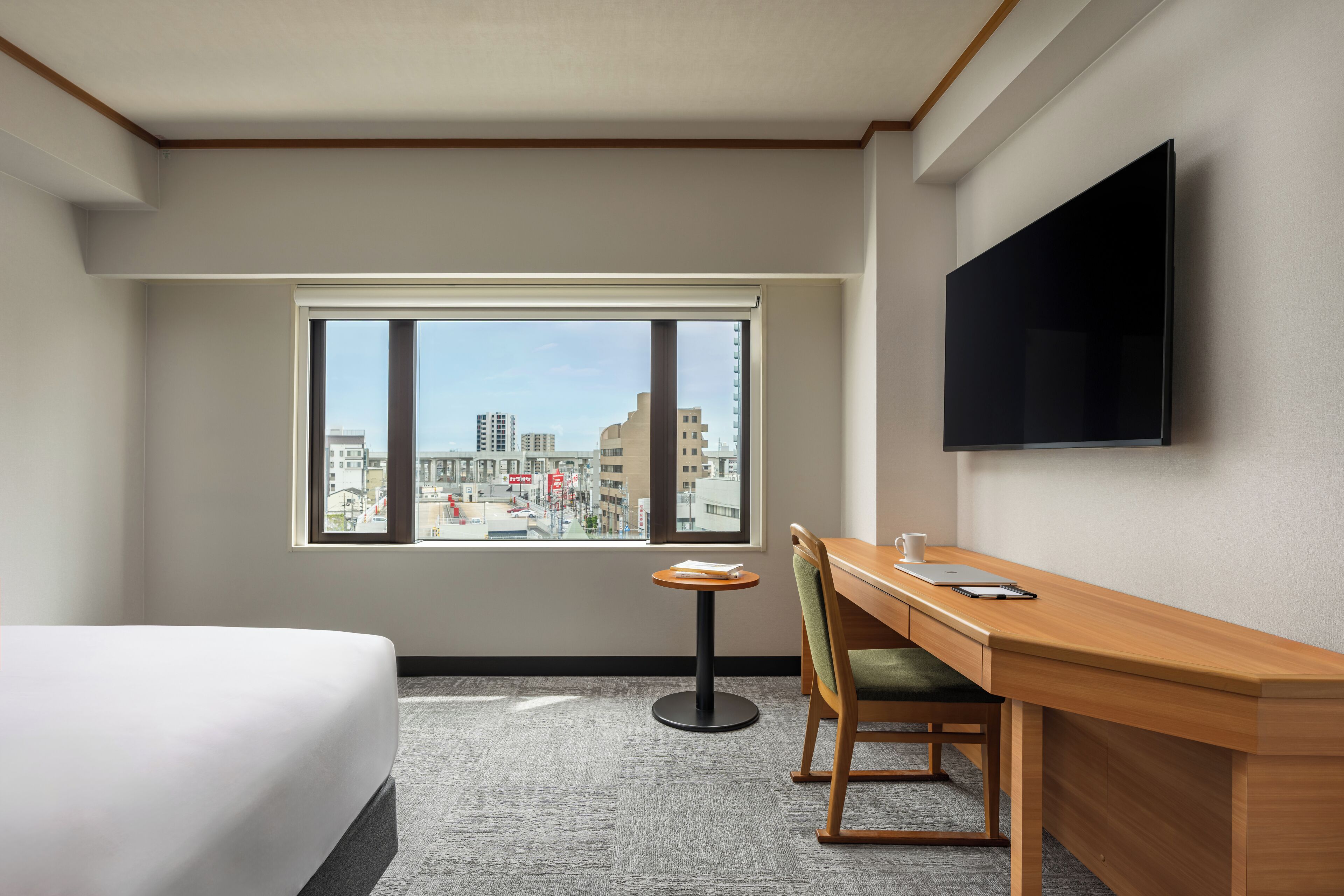 A hotel room with a bed, a desk with a chair, a TV mounted on the wall, and a large window offering a city view.