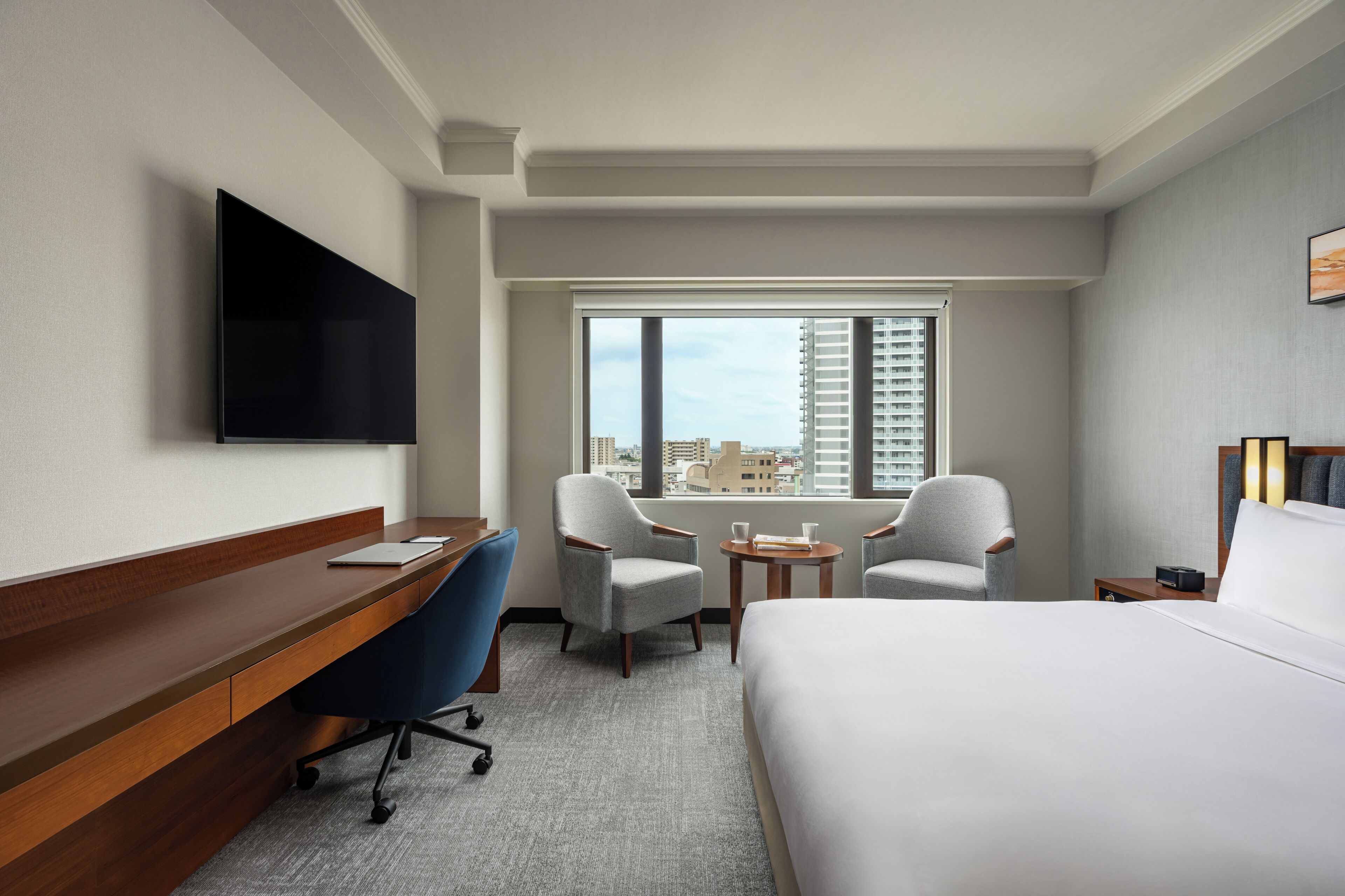Habitación de hotel con cama, escritorio, dos sillones, una mesita, televisor montado en la pared y ventana con vista a la ciudad.