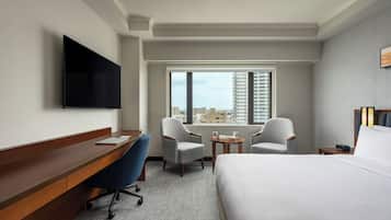 Habitación de hotel con cama, escritorio, dos sillones, una mesita, televisor montado en la pared y ventana con vista a la ciudad.