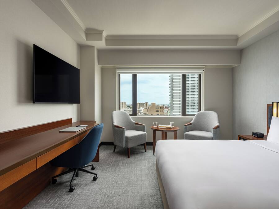 Habitación de hotel con cama, escritorio, dos sillones, una mesita, televisor montado en la pared y ventana con vista a la ciudad.