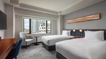 Habitación de hotel con dos camas, un escritorio, una silla y ventana con vista a la ciudad.