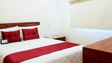 A hotel room with a bed, a wooden headboard, a nightstand with a phone, and a window with blinds.