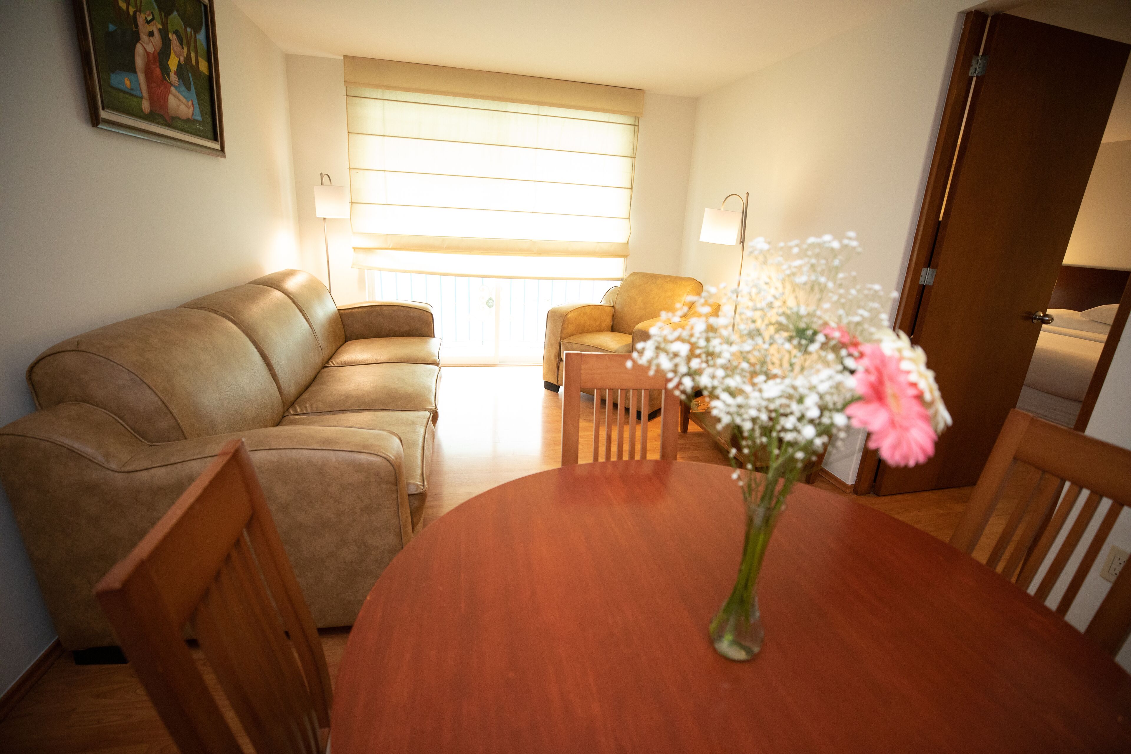 A cozy living room with a brown leather sofa, a wooden dining table with a vase of flowers, and a wooden chair.