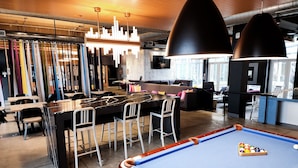 A pool table with a blue felt surface and a set of colorful balls. There are bar stools and a bar area with a counter. The room has a modern design with a wooden ceiling and pendant lighting.