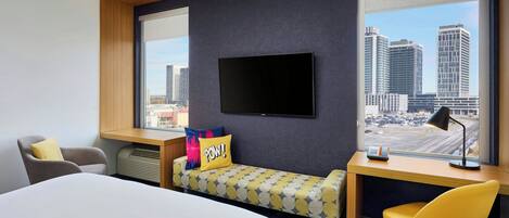 Habitación de hotel con cama, escritorio, silla, televisor y vista a la ciudad.