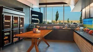 A modern interior with a wooden table, refrigerated beverage dispensers, and a view of parked cars outside.
