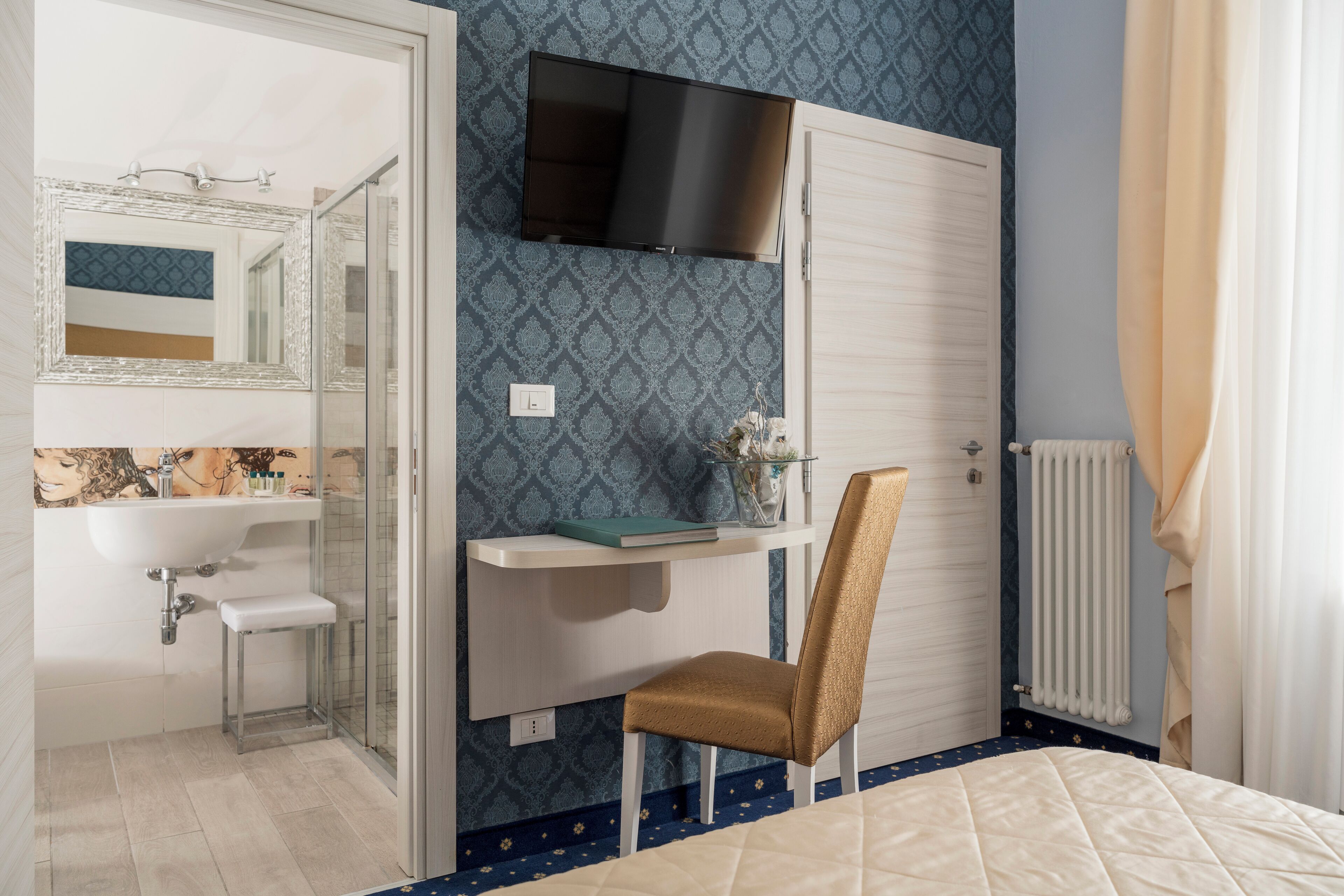 A bedroom with a bed, a desk with a chair, a TV mounted above the desk, a radiator, and a bathroom visible through an open door.